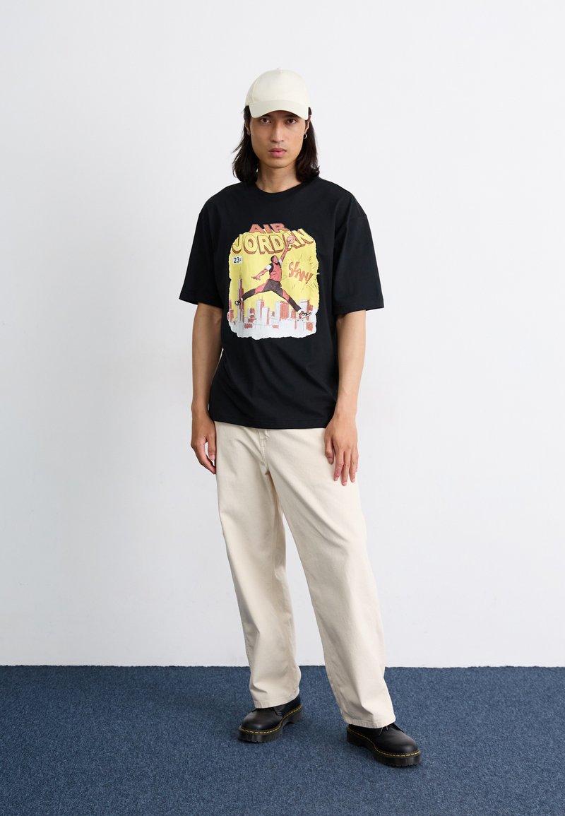 Schwarzes T-Shirt mit einem bunten Grafikdruck einer springenden Figur und Text. Kombiniert mit lockeren, cremefarbenen Hosen und schwarzen Schuhen.