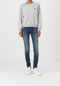 Sudadera gris con un pequeño logo negro, combinada con jeans ajustados azules y zapatillas blancas. Tejido suave con puños y dobladillo acanalados.