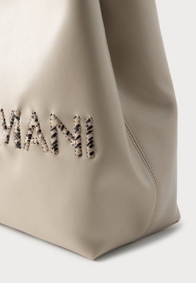 Μπεζ δερμάτινη τσάντα με ανάγλυφη ραφή που σχηματίζει τη λέξη «MANI». Διαθέτει λεία υφή και διακριτικές καμπύλες. Ορατές λεπτομέρειες ραφής στο πλάι.