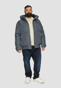 Grijze, geïsoleerde pufferjas met een capuchon, een ritssluiting aan de voorkant en elastische manchetten, gedragen over een lichte trui en donkere spijkerbroeken, gecombineerd met witte sneakers.