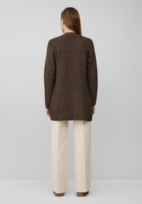 Brauner, gestrickter, langer Cardigan mit einem strukturierten Muster, gestaltet mit einem geraden Schnitt. Kombiniert mit hellen, weit geschnittenen Hosen.