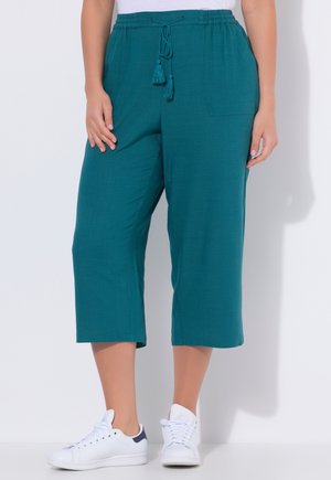 Pantaloni corti turchesi con tasche frontali e coulisse con nappine, indossati con sneakers bianche da una persona in piedi su uno sfondo bianco.