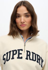 Beige zip-up sweatshirt met marineblauwe "SUPER DRY" tekst. Voorzien van een kraag met een contrasterend blauw ruitpatroon. Zachte textuur.