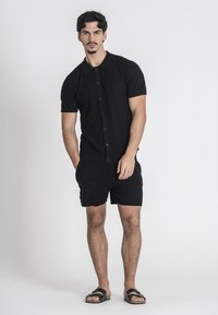 Maglietta nera a maniche corte in maglia con un motivo testurizzato e bottoni, abbinata a pantaloni corti neri e sandali minimalisti.