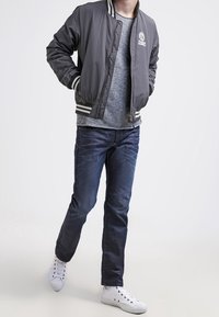 Grå bomberjacka med vita ränder, bärs över en ljusgrå tröja, ihop med mörkblå jeans och vita sneakers, stående mot en neutral bakgrund.