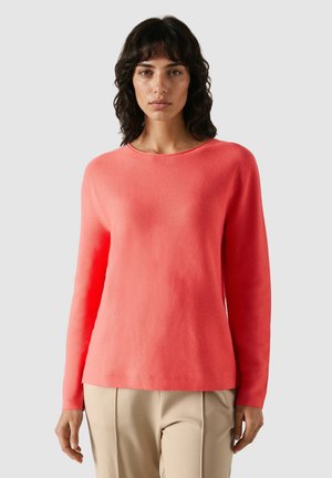 Mujer con cabello oscuro y ondulado, vestida con un suéter coral de manga larga y pantalones beige, posando frente a un fondo liso de color gris claro.