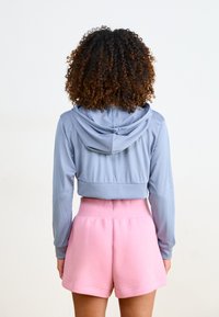 Sudadera azul de malla recortada con capucha, acompañada de pantalones cortos rosa de cintura alta. Textura de tela suave con cintura elástica y detalles en el dobladillo.
