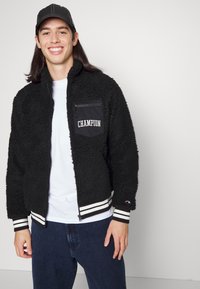 Sort fleecejakke med lynlås på brystlommen og "Champion"-logo. Har stribede manchetter og krave. Sat sammen med en hvid skjorte og mørke jeans.