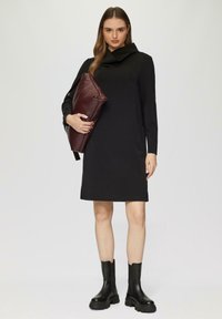 Zwarte lange jurk met een kraag, stevige textuur, tot op de knie. Model houdt een bordeauxkleurige gewatteerde tas vast; draagt zwarte chunky enkellaarzen.
