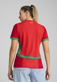 Röd och grön sporttröja med texturerade ärmar, mönstrade accenter och rundad nederkant. Tyget verkar vara lätt och ventilerat.