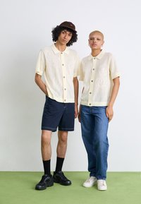 Lysegul, kortermede paisley strikkejakker med knapper, kombinert med marineblå shorts og blå jeans, båret med mørke og lyse joggesko.