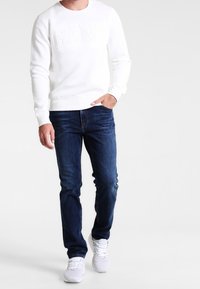 Vit crew-neck sweatshirt med präglad "GANT"-text, kombinerad med mörkblå jeans och vita sneakers. Avslappnad passform, i bomullsblandning.