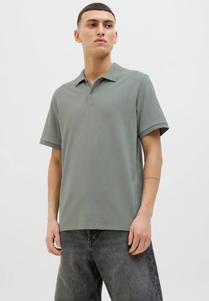 Polo a maniche corte in tessuto verde tenue con colletto, placca a due bottoni e vestibilità rilassata, abbinato a jeans grigio scuro.