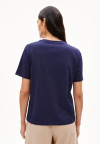 Frau mit langen dunklen Haaren, die sich abwendet, trägt ein schlichtes, navyblaues T-Shirt mit kurzen Ärmeln und beige Hosen vor einem weißen Hintergrund.