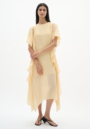 Žena stojaca s prekríženými rukami, oblečená v splývavých bledozelených midi šatách s volánovými rukávmi a čiernych sandáloch s nízkym podpätkom.