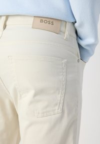 Beige broek met een gladde textuur, voorzien van achterzakken en een lederen label met de opdruk "BOSS" op de tailleband.