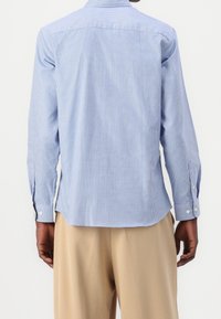 Hombre vistiendo una camisa de manga larga a cuadros azul claro y pantalones beige, visto desde atrás sobre un fondo blanco liso.