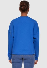 Felpa blu oversize con maniche lunghe, scollatura rotonda e texture morbida. Apparentemente calda e casual, mostrata da dietro con jeans.