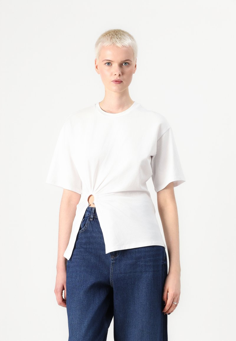 Tricou alb din bumbac cu mânecă scurtă, având un tiv răsucit și un detaliu decupat în formă circulară. Asortat cu blugi din denim albastru cu talie înaltă.
