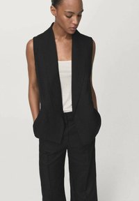 Blazer negro sin mangas con un diseño a medida, que cuenta con dos bolsillos frontales y una textura suave, combinado con una blusa de color claro.