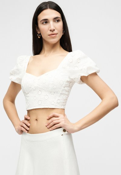 Femme aux longs cheveux foncés portant des boucles d’oreilles anneaux dorés, un crop top blanc à œillets fleuris avec manches bouffantes, et une jupe taille haute blanche.