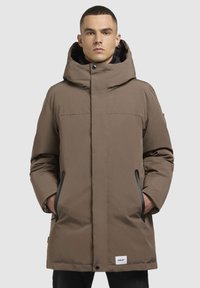Bruine gewatteerde parka met een hoge kraag, grote capuchon, vo pockets en zwarte accenten. Soepele textuur met zichtbare stikseldetails.