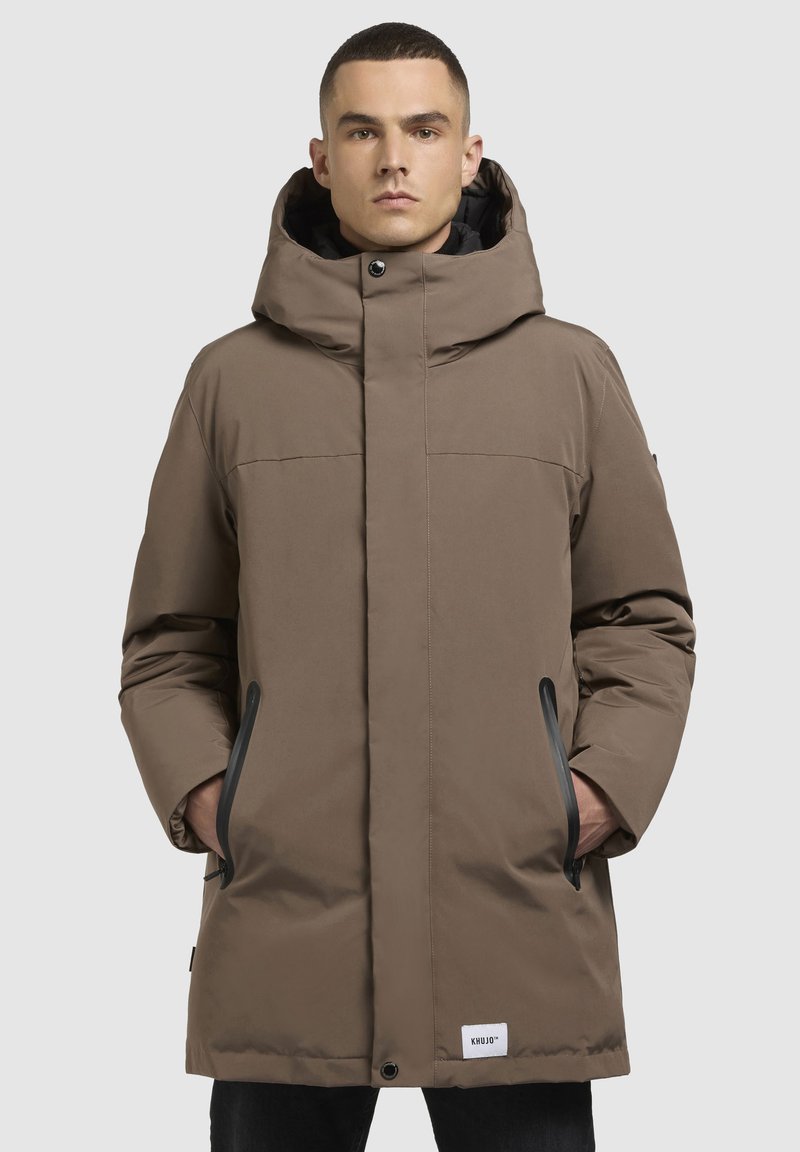 Bruine gewatteerde parka met een hoge kraag, grote capuchon, vo pockets en zwarte accenten. Soepele textuur met zichtbare stikseldetails.