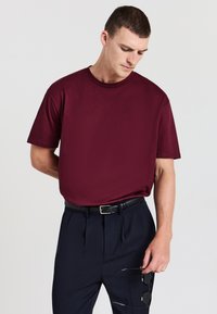 Uomo che indossa una t-shirt bordeaux a maniche corte infilata in pantaloni neri a pieghe con una cintura nera, mentre tiene occhiali da sole neri in una mano.