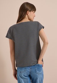 Femme aux longs cheveux bruns portant un top à manches courtes rayé noir et blanc et un jean bleu, debout avec une main dans sa poche.