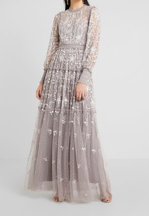 Femme portant une longue robe lavande transparente avec des broderies florales blanches, manches longues, taille froncée et jupe à volants, debout devant un fond uni.
