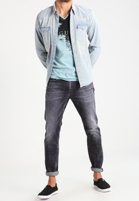 Camisa de mezclilla azul claro sobre una camiseta gráfica negra y jeans gris oscuro, combinados con zapatos negros sin cordones. Piernas ligeramente entalladas, puños doblados.