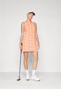 Golf ruha élénk rózsaszín és narancssárga kockás mintával, gallérral, ujjatlan kialakítással, golfütővel és fehér sapkával, fehér cipővel párosítva.