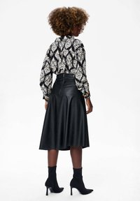 Vrouw met krullend haar, gekleed in een zwart-witte blouse met patroon, een zwarte leren midi-skirt met een ritssluiting aan de achterkant, en zwarte leren enkellaarsjes met hak.