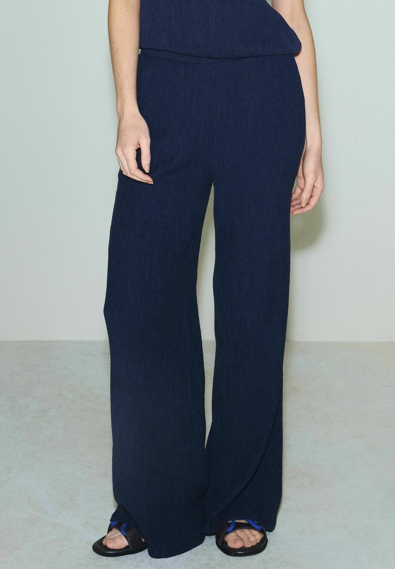 Femme portant un pantalon large ample bleu marine et des sandales noires à bout ouvert, debout sur un sol gris clair devant un mur uni.