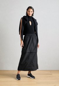 Black knit top with a keyhole detail, layered black long skirt with pockets, and black slip-on shoes with a textured finish.