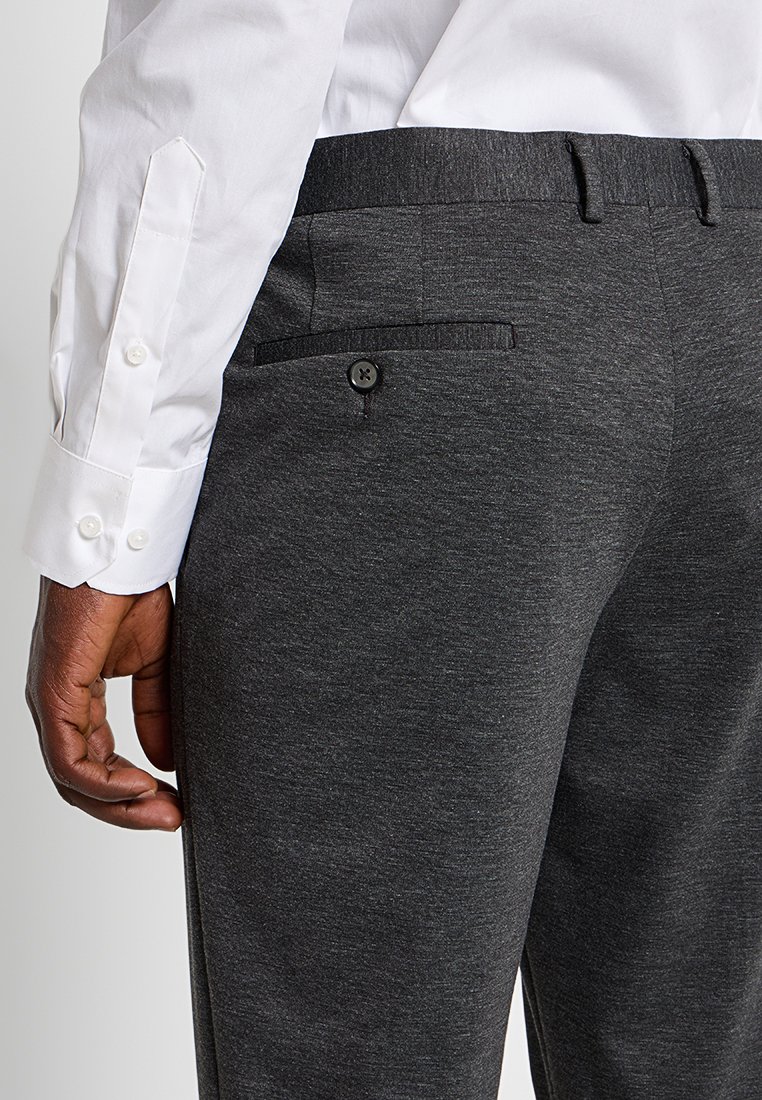 Pantalones grises con una textura suave, que presentan un bolsillo trasero con botón y un frente plano. Llevados con una camisa blanca de manga larga.