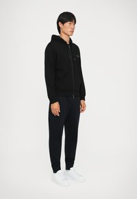 Svart zip-up hettegenser laget av myk stoff, med snøring på hetta. Kombinert med marineblå joggebukser og hvite sneakers for en avslappet look.