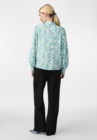 Blusa azzurra fluida con un motivo floreale nei toni verdi e blu, caratterizzata da maniche a palloncino e una vestibilità rilassata, abbinata a pantaloni neri.