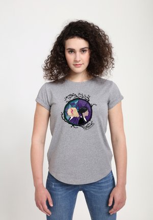 Femme aux cheveux bouclés portant un t-shirt gris affichant deux femmes illustrées, dos à dos, à l'intérieur d'un cadre circulaire avec des branches et des toiles d'araignée.