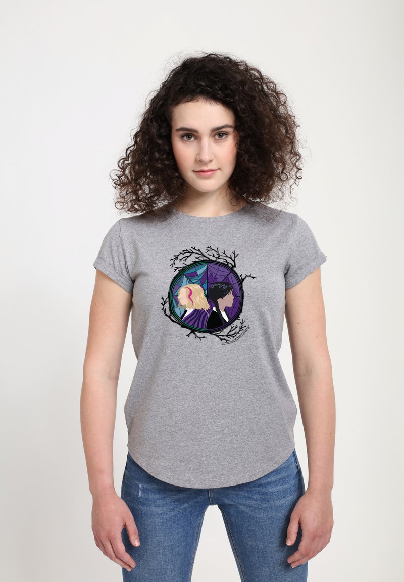 Kvinde med krøllet hår bærer en grå t-shirt, der viser to illustrerede kvinder ryg-til-ryg inde i en cirkulær ramme med grene og edderkoppespidser.