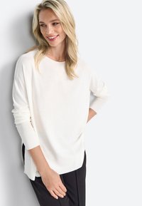 Pull léger blanc avec des manches longues, des poignets côtelés et des fentes sur les côtés, associé à un pantalon noir. Texture lisse et coupe décontractée.