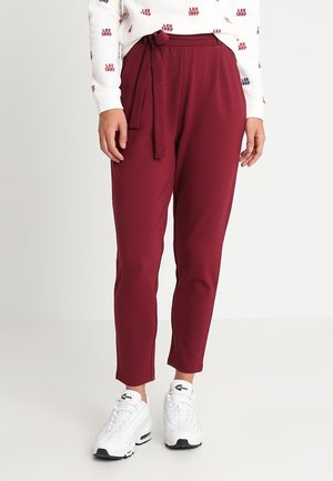 Mujer con pantalones burdeos de talle alto con cinturón en la cintura y zapatillas blancas, combinados con una camiseta blanca de manga larga.