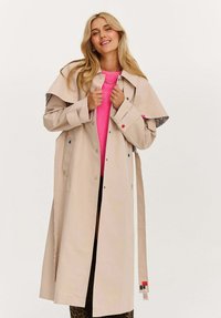 Blonďatá žena se usmívá, má na sobě dlouhý béžový trenčkot přes jasně růžový svetr a leopardí kalhoty, stojí na neutrálním pozadí.