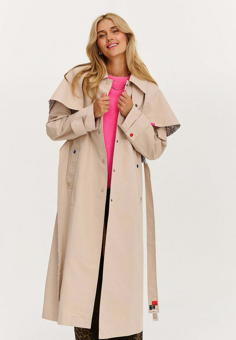 Blonďatá žena se usmívá, má na sobě dlouhý béžový trenčkot přes jasně růžový svetr a leopardí kalhoty, stojí na neutrálním pozadí.