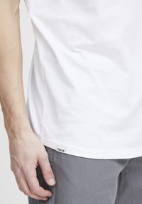 Hvit bomulls T-skjorte med korte ermer, som har en slim fit og liten etikett på nederste søm. Grå bukser delvis synlige.