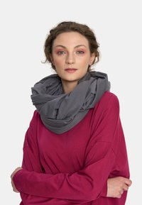 Kobieta z kręconymi włosami, nosząca dużą szaro-skalową chustę i długą, różową bluzkę z długim rękawem, stoi z założonymi na piersiach rękami na jednolitym tle.