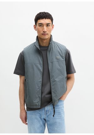 Junger Mann, der eine ärmellose graue Jacke mit Reißverschluss über einem dunkelgrauen T-Shirt und hellblauen Jeans trägt, vor einem schlichten weißen Hintergrund stehend.