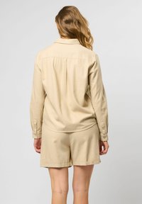 Beige overhemd met lange mouwen en opgerolde manchetten, gecombineerd met bijpassende shorts. Zacht materiaal, minimalistisch ontwerp en een getailleerde pasvorm.