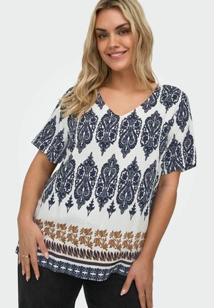 Uśmiechnięta kobieta w białej bluzce z krótkim rękawem, ozdobionej wzorami kwiatowymi i geometrycznymi w granatowym i brązowym kolorze, stoi na tle o jednolitym kolorze.