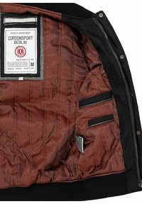 Schwarze Jacke mit einem gesteppten braunen Futter, ausgestattet mit einem Etikett, zwei Reißverschlusstaschen und gerippten Bündchen. Gewebter Stoff mit gepolsterten Details.